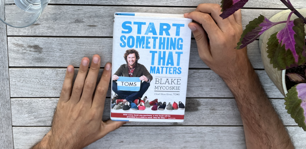 Man holding the front cover of Start Something That Matters by Blake Mycoskie on a table.